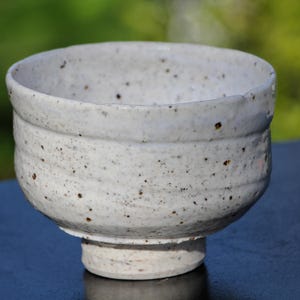 May include: A handmade, off-white ceramic bowl with a speckled texture. The bowl has a wide, rounded shape with a slightly textured rim and a sturdy, cylindrical base. The exterior features small, dark brown speckles.