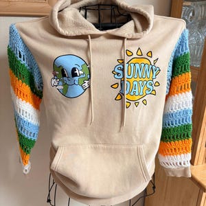 May include: A beige hooded sweatshirt featuring a front graphic of a cartoon Earth and a yellow sun with the text "SUNNY DAYS". The long sleeves are made of multi-colored crocheted panels in green, orange, white, and light blue, with a front pocket.
