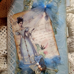 May include: A handmade journal with a vintage aesthetic. The cover is light blue with a floral pattern and a lace trim. It features a printed image of a woman in a blue dress, a blue tulle bow, and a decorative butterfly.