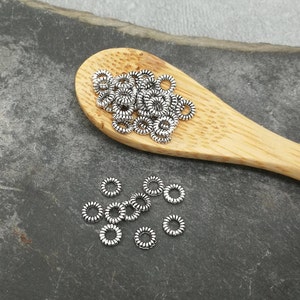 Op de afbeelding: Een stapel zilveren springringen op een houten lepel. De springringen zijn klein en hebben een licht getextureerd oppervlak. Er liggen ook een paar springringen verspreid op een grijs oppervlak.