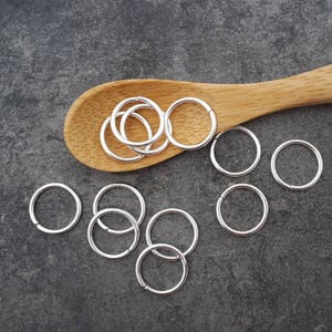 May include: A pile of silver jump rings on a wooden spoon, with additional jump rings scattered on a gray surface.