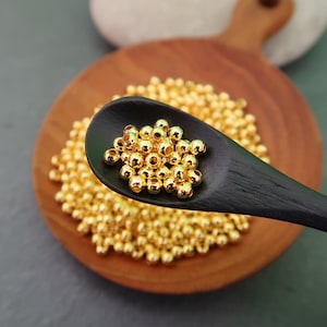 May include: A wooden spoon filled with gold-colored round beads. The beads are arranged in a pile on a wooden surface.