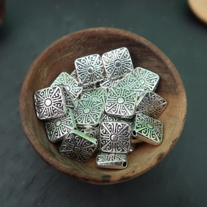 May include: A bowl filled with silver metal square beads with intricate designs. The beads have a raised center design and are perfect for jewelry making.