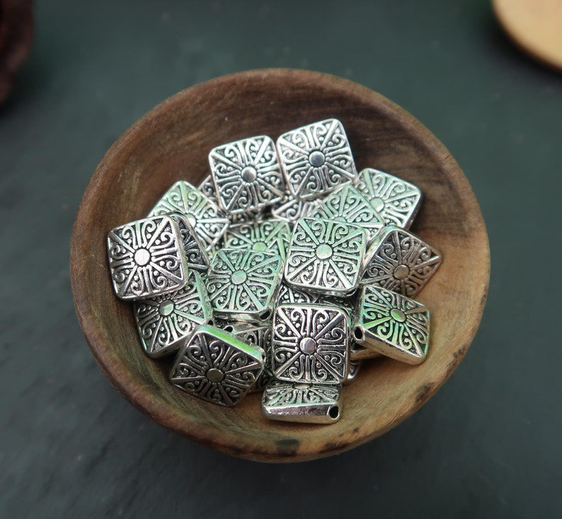 May include: A bowl filled with silver metal square beads with intricate designs. The beads have a raised center design and are perfect for jewelry making.