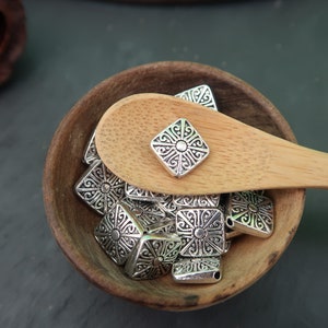 May include: A wooden bowl filled with silver-toned metal charms. The charms are square-shaped with an intricate design. A wooden spoon with a single charm on top is resting on the edge of the bowl.