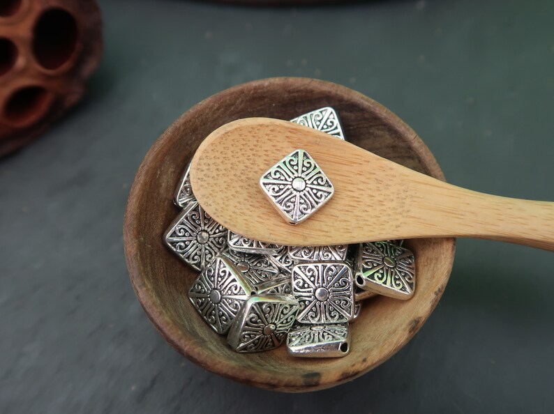 May include: A wooden bowl filled with silver-toned metal charms. The charms are square-shaped with an intricate design. A wooden spoon with a single charm on top is resting on the edge of the bowl.