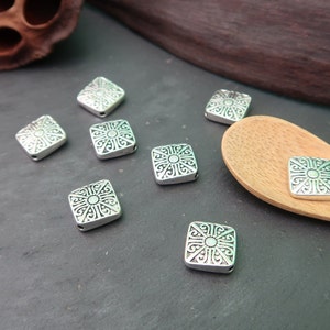 May include: Silver square beads with an intricate design. The beads have a hole in the center for stringing.
