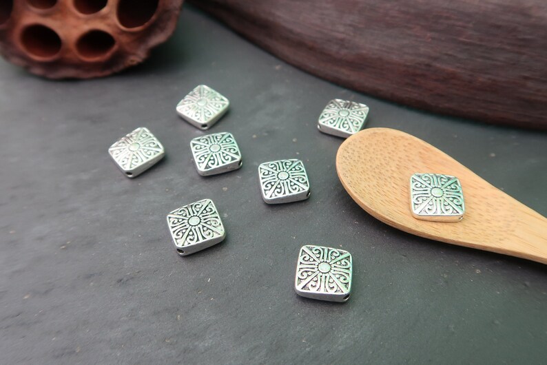 May include: Silver square beads with an intricate design. The beads have a hole in the center for stringing.