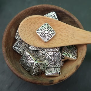 May include: A wooden bowl filled with silver metal charms. The charms are square with a decorative design in the center. The bowl is sitting on a dark green surface.