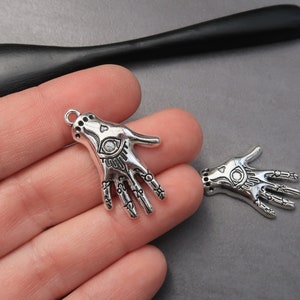 May include: Two silver charm pendants in the shape of a hand with an eye in the palm. The hands are detailed with lines and symbols.