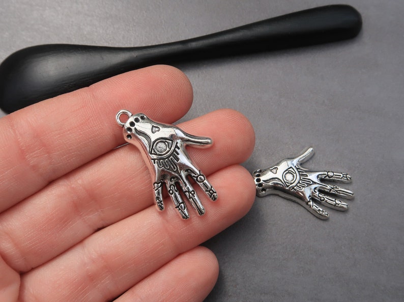 May include: Two silver charm pendants in the shape of a hand with an eye in the palm. The hands are detailed with lines and symbols.