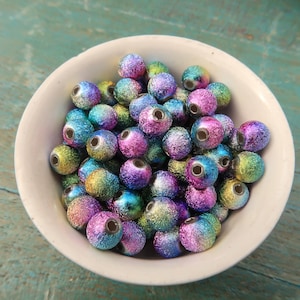 May include: A bowl filled with iridescent round beads. The beads are a mix of blue, green, purple, and gold colors. The beads have a textured surface and a metallic sheen.