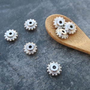 Op de afbeelding: Zilveren metalen kralen met een decoratief ontwerp. De kralen zijn rond met een verhoogd midden en een gegolfde rand. De kralen liggen op een houten lepel en een grijs oppervlak.