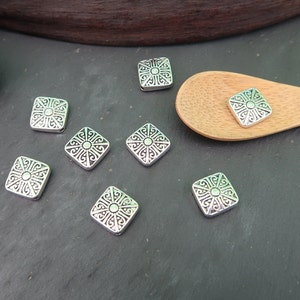 May include: Eight silver square-shaped metal beads with an intricate design. The beads have a raised center and a decorative border.