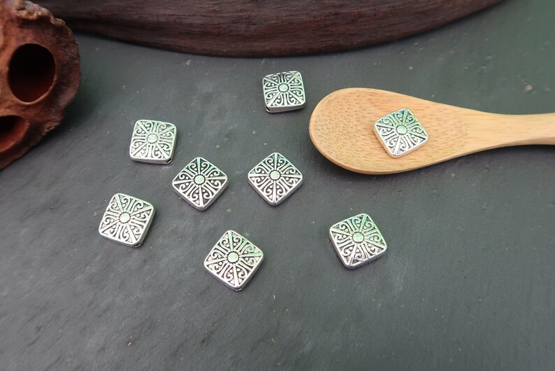 May include: Eight silver square-shaped metal beads with an intricate design. The beads have a raised center and a decorative border.