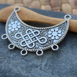 May include: Silver-toned metal crescent moon pendant with a decorative knot design and floral accents. The pendant has five jump rings for attaching chains or cords.