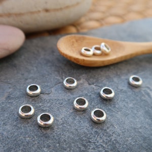 May include: A collection of small, silver-colored metal beads with a rounded, donut-like shape. Several beads are scattered on a dark gray surface, with a few more resting in a wooden spoon. The beads have a smooth, shiny finish.
