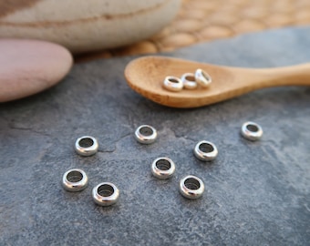 May include: A collection of small, silver-colored metal beads with a rounded, donut-like shape. Several beads are scattered on a dark gray surface, with a few more resting in a wooden spoon. The beads have a smooth, shiny finish.