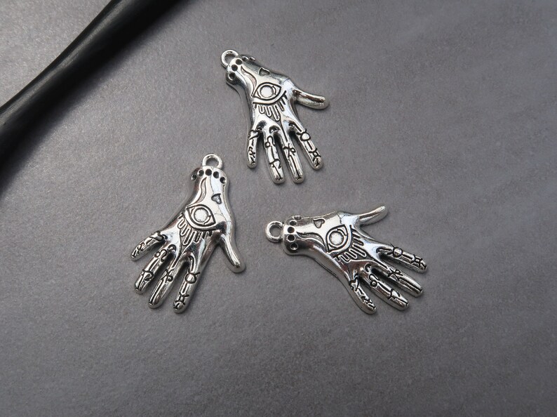 May include: Three silver charm pendants in the shape of a hand with an eye in the palm. The charms are detailed with lines and patterns.