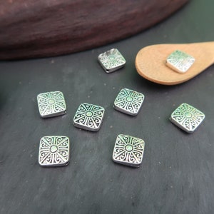 May include: Silver square beads with an intricate design. The beads have a raised, ornate pattern in the center and around the edges.