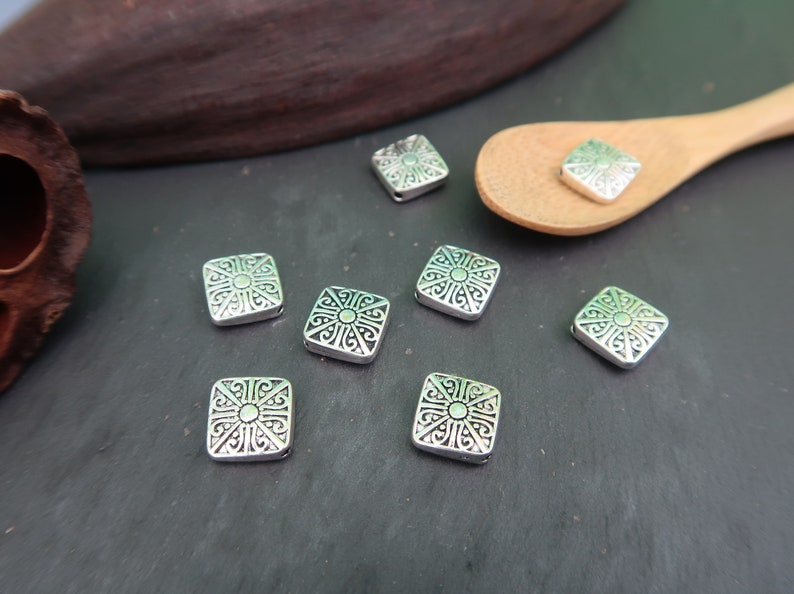 May include: Silver square beads with an intricate design. The beads have a raised, ornate pattern in the center and around the edges.