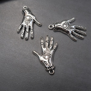 May include: Three silver charm pendants in the shape of a hand with an eye in the palm. Each hand has intricate details and a loop at the top for attaching to a chain or bracelet.
