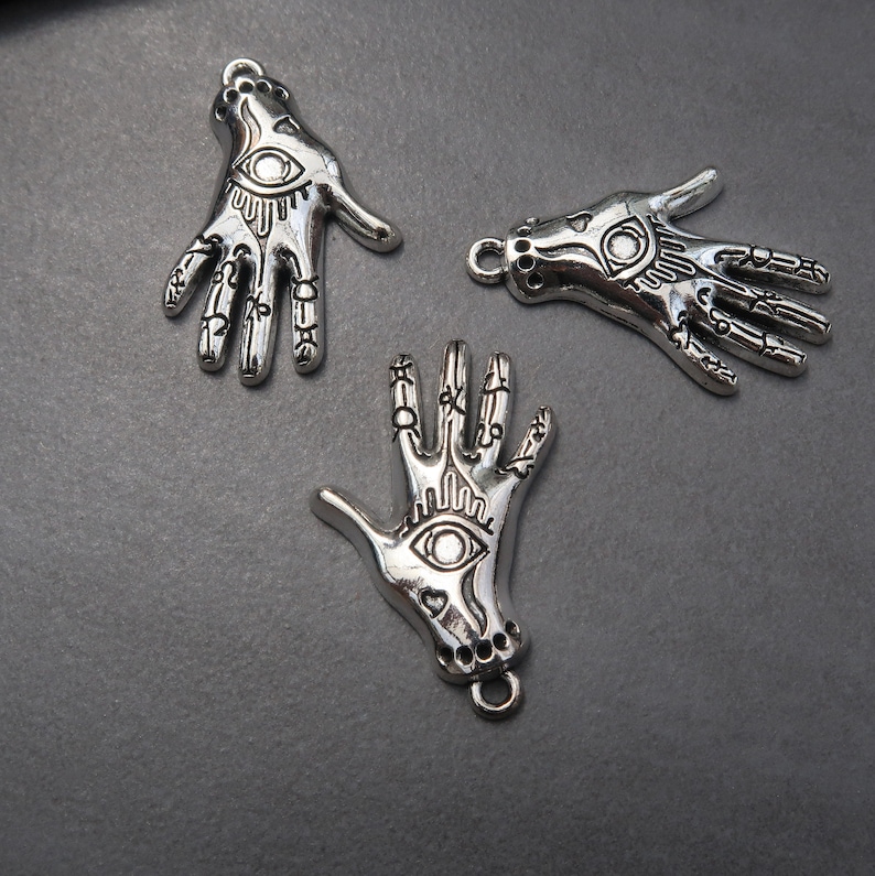 May include: Three silver charm pendants in the shape of a hand with an eye in the palm. Each hand has intricate details and a loop at the top for attaching to a chain or bracelet.