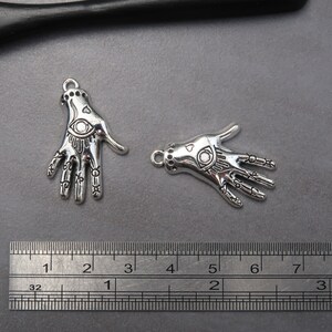 May include: Two silver charm pendants in the shape of a hand with an eye design. The charms are approximately 2 inches long and are shown next to a ruler for size reference.