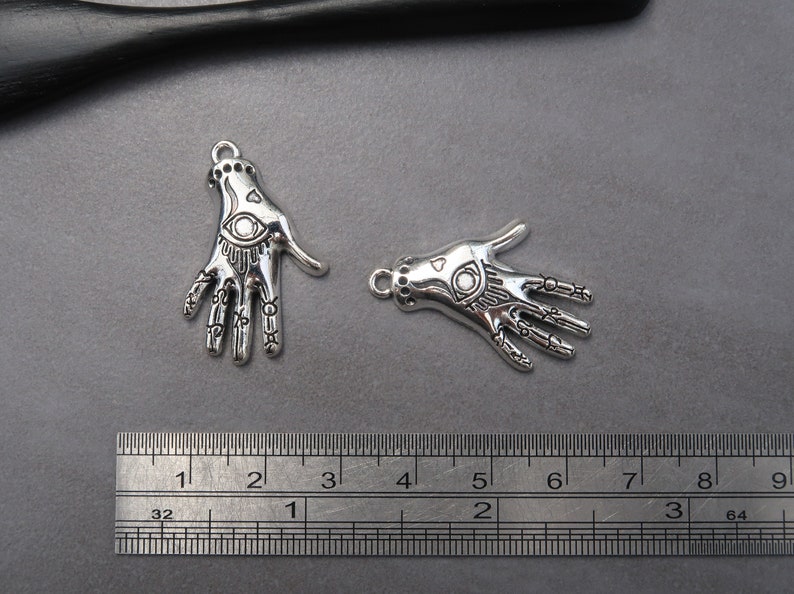 May include: Two silver charm pendants in the shape of a hand with an eye design. The charms are approximately 2 inches long and are shown next to a ruler for size reference.