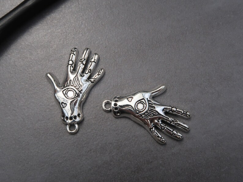 May include: Two silver charm pendants in the shape of a hand with an eye design. The charms have a detailed design and are perfect for jewelry making.