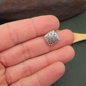 May include: Silver square charm with an intricate floral design.