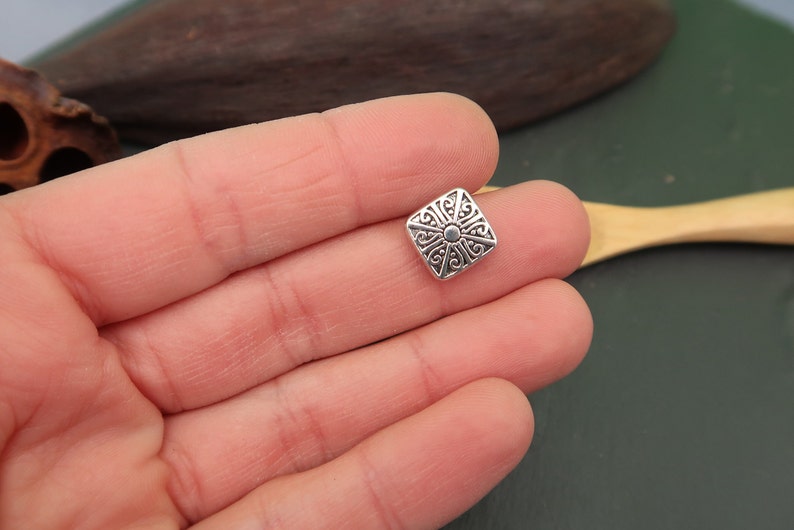 May include: Silver square charm with an intricate floral design.