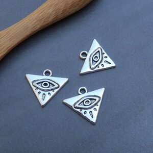 Peut inclure: Trois breloques triangulaires argentées avec un motif d'œil. Chaque breloque a une boucle en haut pour l'attacher à des bijoux ou d'autres objets artisanaux.
