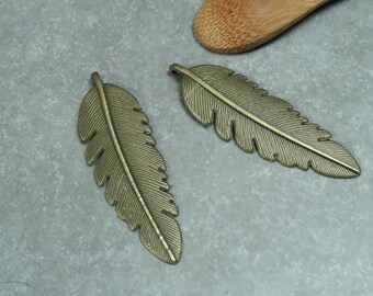 Puede incluir: Dos abalorios de plumas de metal de color dorado antiguo. Las plumas tienen un diseño detallado y texturizado.