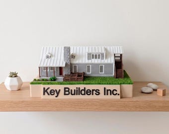 May include: A miniature model of a gray house with a white metal roof, a stone chimney, and a porch, mounted on a green base. The front of the base features the black text Key Builders Inc. in a clean, sans-serif font, displayed on a light tan panel.