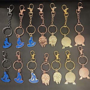 Puede incluir: Colección de llaveros con varios dijes. Los dijes incluyen un sombrero de mago azul, una nave espacial y una esfera dorada. Los llaveros tienen acabados plateados, dorados y oro rosa. Cada llavero tiene un cierre y un anillo.