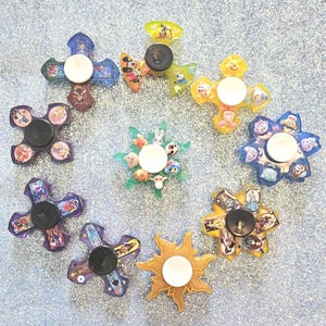 May include: A collection of colorful fidget spinners with various designs, including cartoon characters and floral patterns. Each spinner has a central button and three or four arms. The spinners are arranged on a sparkly, light blue surface.
