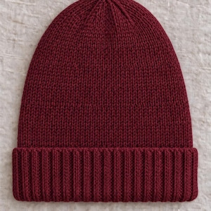 May include: A deep burgundy knit beanie featuring a classic stockinette stitch pattern on the main body and a thick, ribbed cuff at the base. The hat is constructed from a solid-colored yarn with a consistent texture throughout the entire piece.