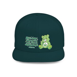 Peut inclure: Une casquette de baseball bleu sarcelle foncé avec un motif représentant un ours vert clair tenant une cigarette, avec une feuille de cannabis sur le ventre. Le texte indique : Life's better when you're soft, slow & slightly stoned. Fuzzy Baby.