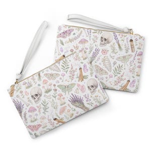 May include: Two white zippered wristlet clutches with a repeating pattern of skulls, butterflies, mushrooms, and lavender sprigs. The clutches have a gold zip and a white wrist strap.