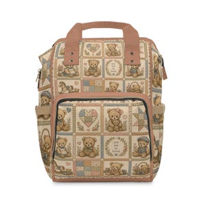 Puede incluir: Una mochila con un estampado estilo patchwork de ositos de peluche, corazones y estrellas en tonos tierra apagados. La bolsa tiene correas y ribetes de color rosa empolvado, un bolsillo frontal con cremallera y el texto you are so loved.
