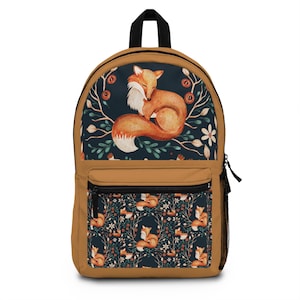 May include: A tan backpack with a black zipper and straps. The top panel features a watercolor illustration of a curled-up fox surrounded by floral elements on a dark blue background. The front pocket has a repeating pattern of foxes and foliage.