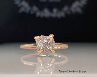 Könnte beinhalten: Ein Ring aus Roségold mit einem rohen Diamanten in der Mitte. Der Diamant ist hellgrau und hat eine raue, natürliche Textur. Der Ring ist ein schlichtes Band mit polierter Oberfläche.
