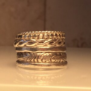 May include: A stack of five silver rings with different textured designs. The top ring has a row of small beads, the second ring has a braided design, the third ring is plain, the fourth ring has a floral design, and the bottom ring is plain.