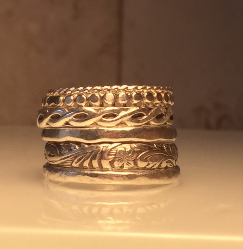 May include: A stack of five silver rings with different textured designs. The top ring has a row of small beads, the second ring has a braided design, the third ring is plain, the fourth ring has a floral design, and the bottom ring is plain.