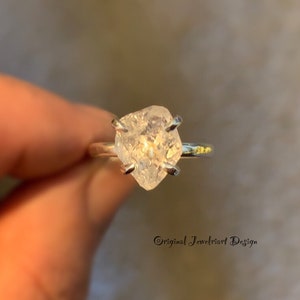 May include: A silver ring with a clear, faceted, rough-cut gemstone. The gemstone is set in a prong setting. The ring is on a finger.