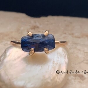 May include: A gold ring with a blue gemstone set in a prong setting. The ring is on a white background.