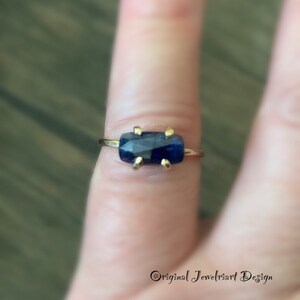May include: A gold ring with a dark blue rectangular gemstone. The ring is on a finger.
