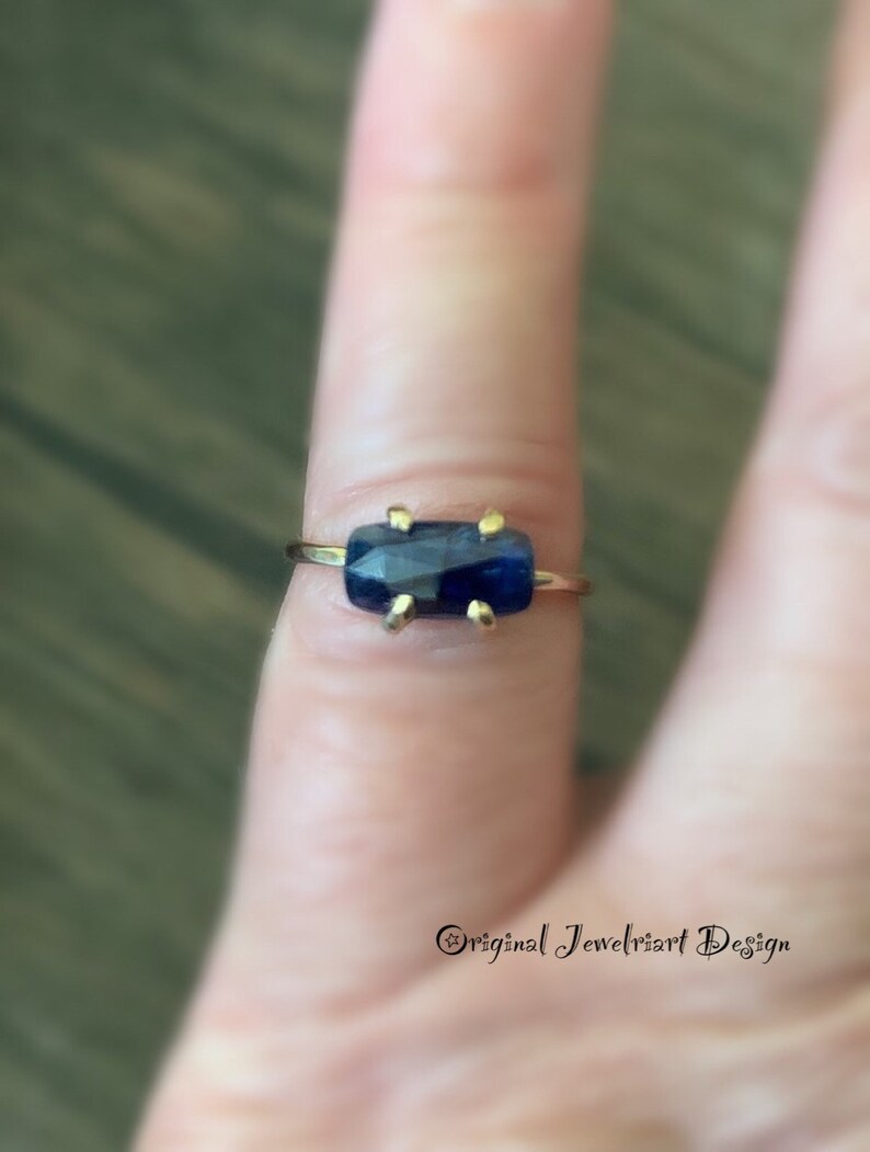 May include: A gold ring with a dark blue rectangular gemstone. The ring is on a finger.