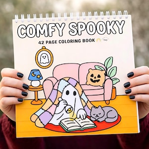 May include: A spiral-bound coloring book titled "COMFY SPOOKY" with a 42-page count. The cover features a cartoon illustration of a ghost under a blanket, reading a book, with a cat, pumpkin, and sofa. The background is yellow.